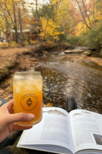 Hand holding a Vacayzy glass of apple cider by a creekside cabin in Blue Ridge, Georgia, with fall leaves and a book in the background.