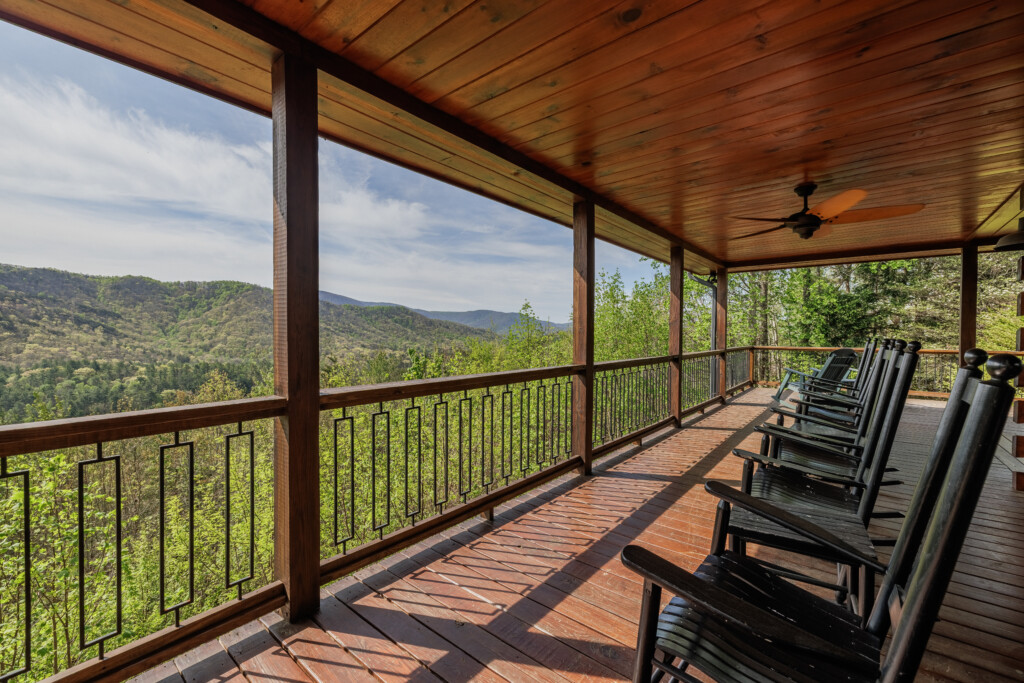 Spacious covered deck at Above the Clouds cabin in Blue Ridge, GA, featuring a cushioned outdoor sectional, teak coffee table, ceiling fan, and rocking chairs — all facing open North Georgia mountain views through black iron railings and towering pines. An outdoor living room built for long, unhurried afternoons.