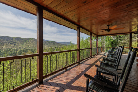 Spacious covered deck at Above the Clouds cabin in Blue Ridge, GA, featuring a cushioned outdoor sectional, teak coffee table, ceiling fan, and rocking chairs — all facing open North Georgia mountain views through black iron railings and towering pines. An outdoor living room built for long, unhurried afternoons.